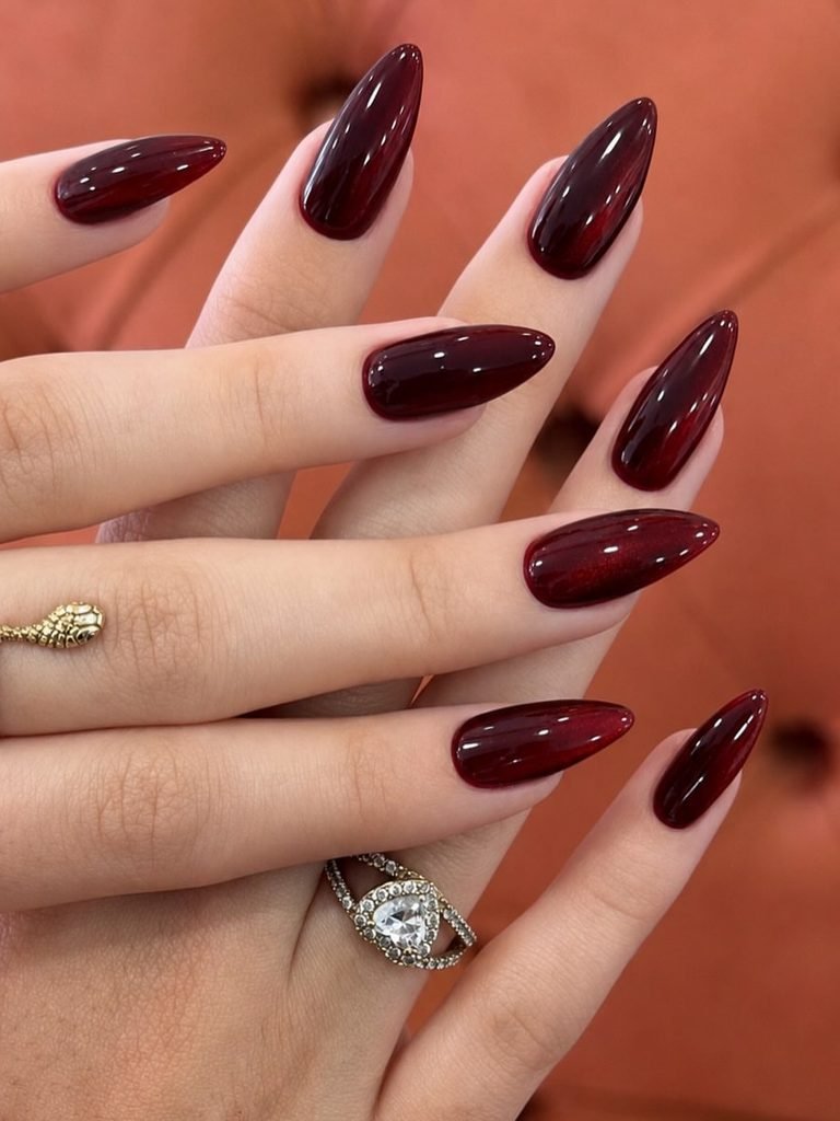 glossy deep burgundy red almond nails styled with gold rhinestone rings