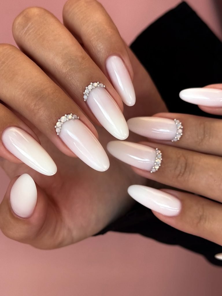 milky white almond nails styled with delicate rhinestone embellishments near the cuticles