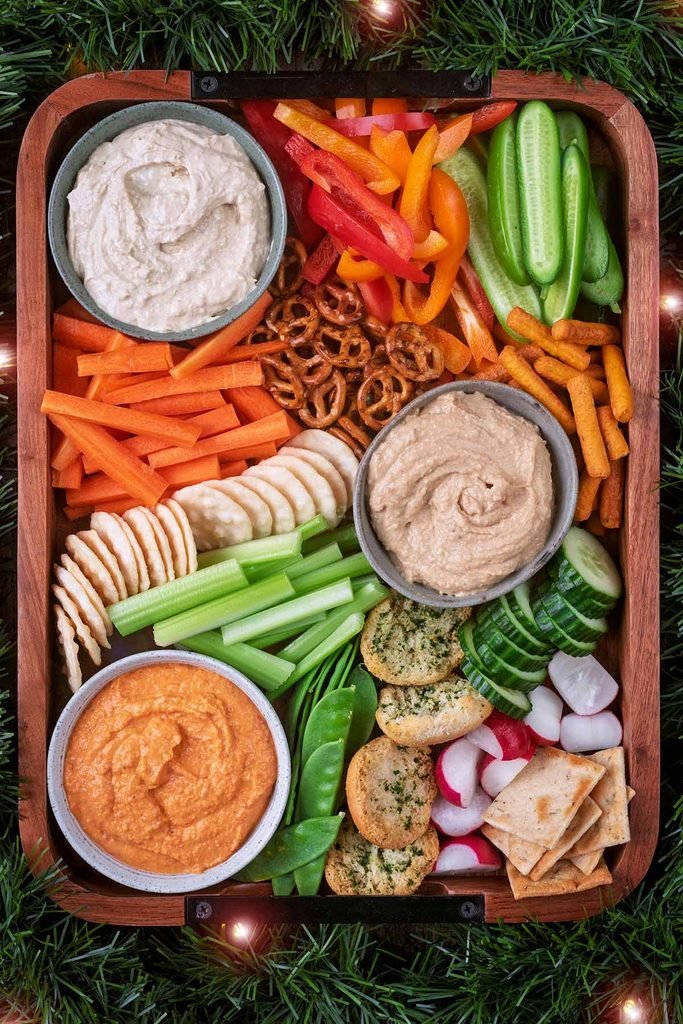 wooden tray platter arranged with varied hummus dips, fresh vegetables, crackers, and pretzels