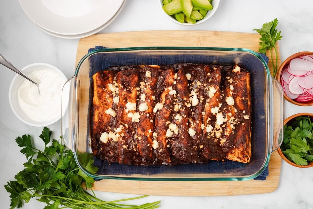 baked bean enchiladas topped with dark sauce and crumbled cheese in a glass dish   