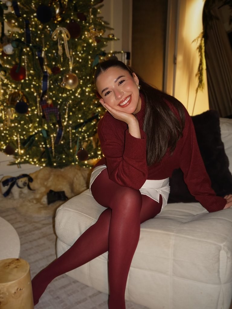 smiling woman wearing a red sweater and tights sitting by a lit christmas tree   
