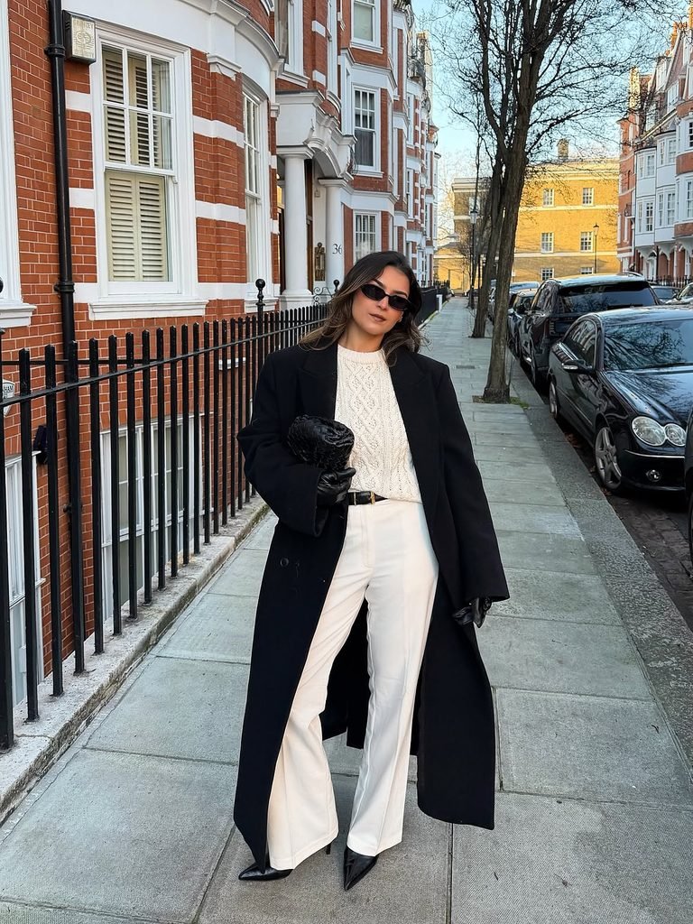 woman wearing a long black coat, white cable knit sweater, and white trousers