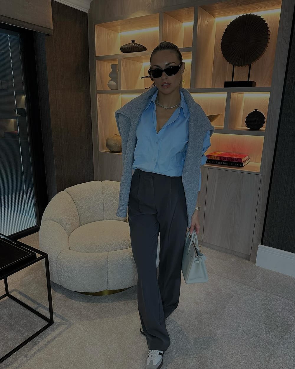 Pale-blue button-down with dark trousers, a gray sweater draped over the shoulders, gray sneakers and a light gray handbag.