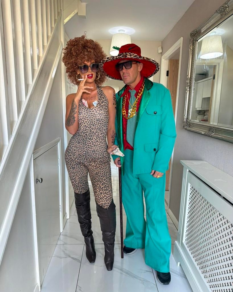 man and woman in colorful pimp costumes posing indoors with gold chains
