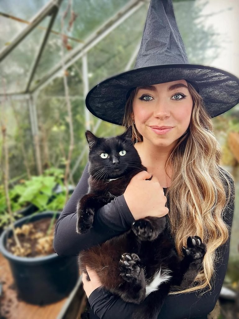 woman with blonde hair wearing a black witch hat while holding a black cat