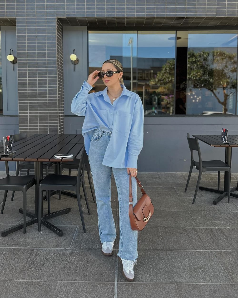 Pale-blue button-down half-tucked into light-wash jeans with gray-and-white sneakers, dark sunglasses and a brown shoulder bag.